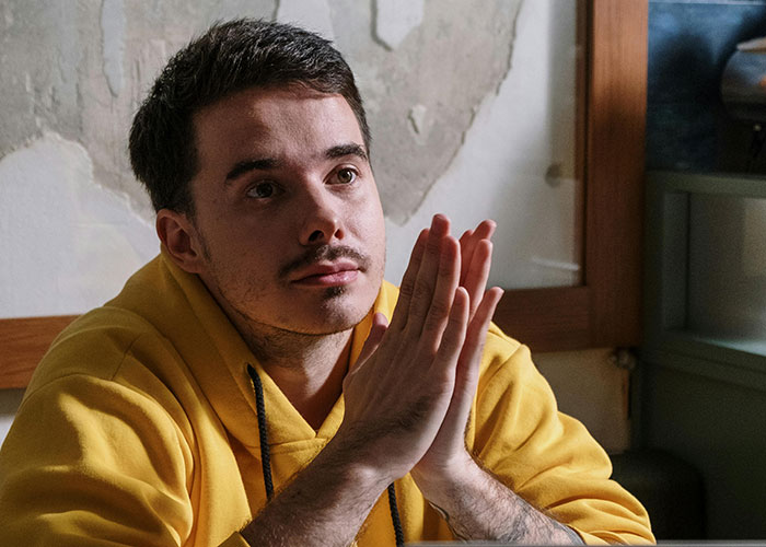Young man in a yellow hoodie thoughtfully clasping hands, illustrating guy asks ex to move out after breakup concern.