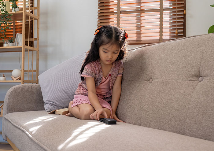 Young girl sitting alone on a couch looking at a remote, depicting a child left alone without babysitting in an emergency.