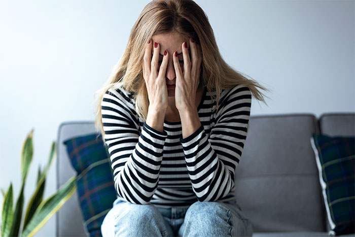 Woman with long blonde hair in striped shirt sitting on couch, questioning marriage after husband falls in love with AI girlfriend.