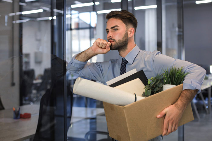 Man holding box of personal items, looking thoughtful and concerned after becoming jobless in an office setting.