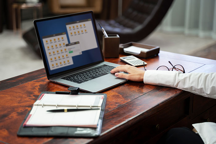Worker at desk using laptop with documents and glasses nearby, appearing annoyed while avoiding birthday cake celebration.