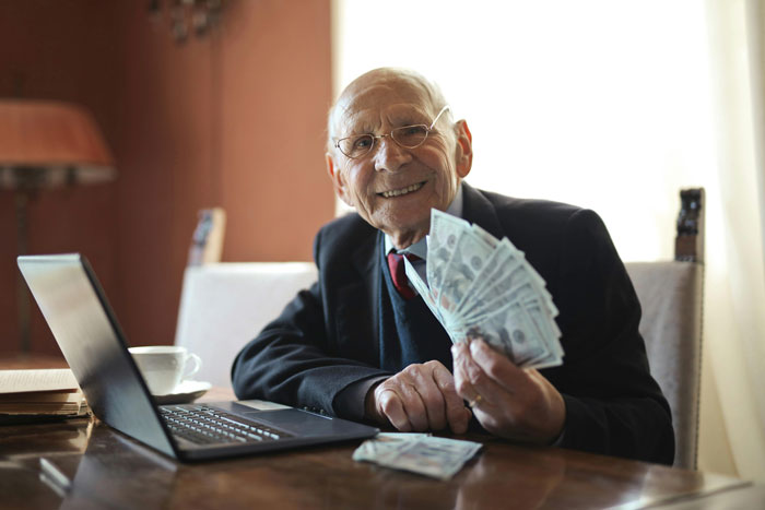 Elderly wealthy grandpa smiling and holding cash while sitting at a table with a laptop, celebrating college gift.